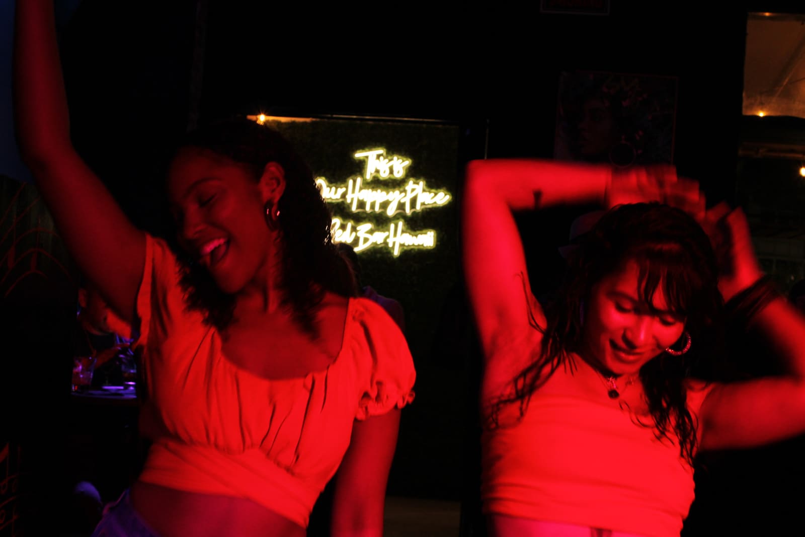 Two guests dancing under red light at Red Bar Hawaii, below the neon sign that reads “This is our happy place”